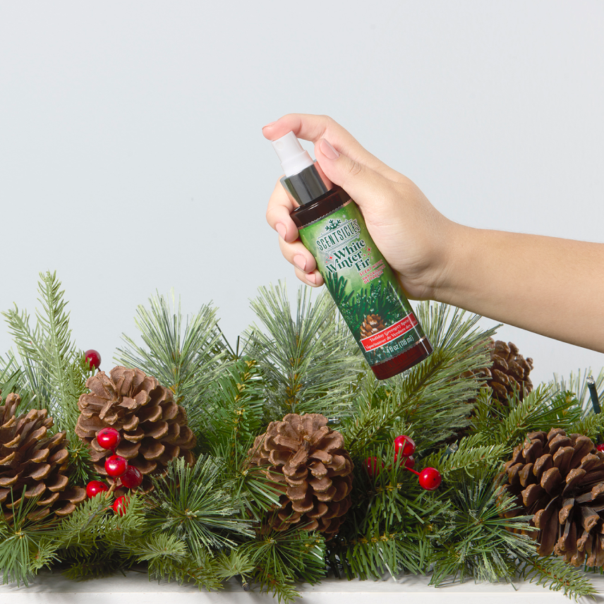 Hand holding Scentsicles White Winter Fir Scented Spray, applying fresh pine fragrance to a holiday wreath with pinecones and berries.