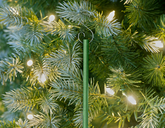 Green scented ornament stick hanging on a Christmas tree, illuminated by warm, glowing string lights, nestled among lush green branches.