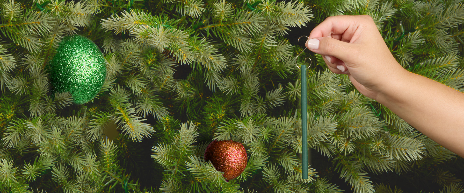 A hand hangs a green scented ornament stick on a Christmas tree, surrounded by green and gold glittering ornaments, blending into the lush tree branches.