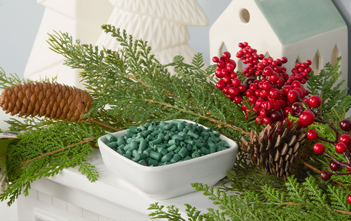 Holiday-themed arrangement featuring a white square dish filled with green scented ornament sticks, surrounded by pinecones, red berries, and evergreen foliage, set against a backdrop of white holiday decorations.