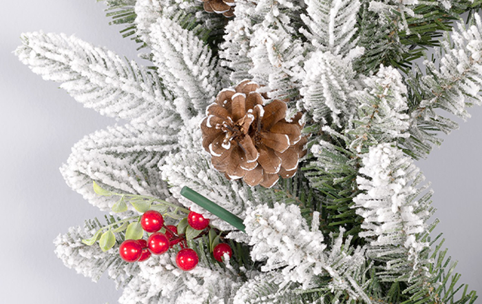 Snow-dusted Christmas tree branch adorned with a pine cone, red berries, and a green scented ornament stick nestled among the frosted foliage.