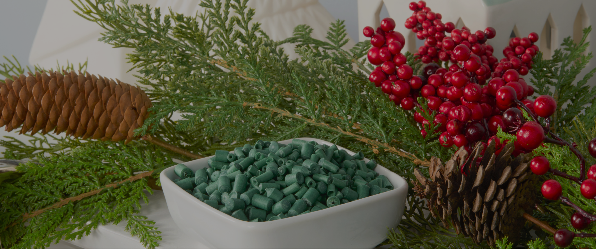 Festive holiday display featuring a pinecone, red berries, and green foliage, complemented by a bowl filled with green scented ornament sticks, set against a backdrop of white holiday decorations.