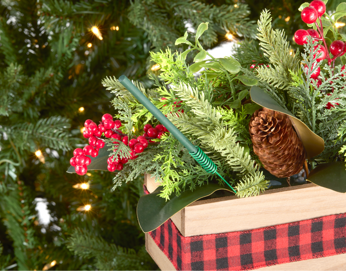 Festive holiday arrangement featuring red berries, green foliage, and a pine cone, accented by a green scented ornament stick, all set in a rustic wooden box with a plaid ribbon, with a Christmas tree in the background.